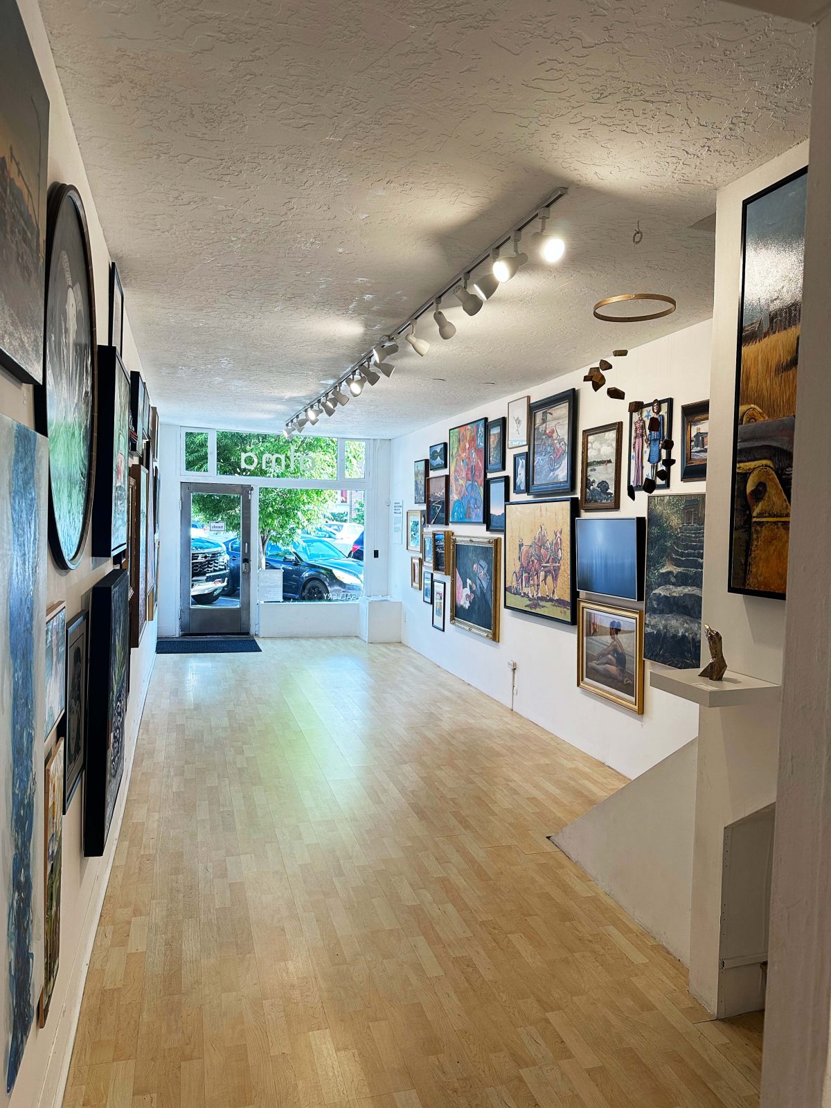 A view down a narrow art gallery lined with densely packed paintings and mixed-media works on both walls, leading toward the entrance with large front windows and light streaming in.