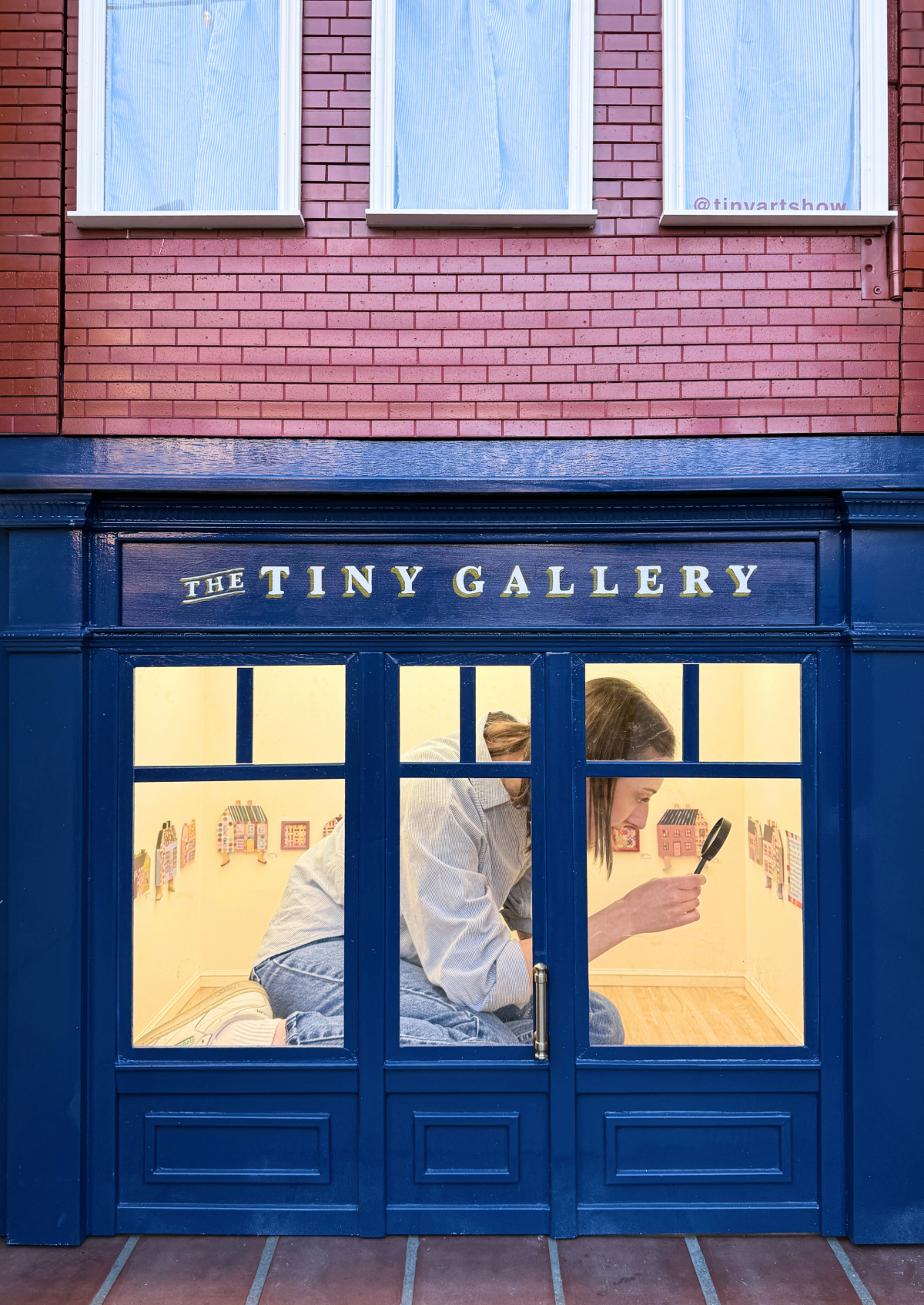 Exterior view of The Tiny Gallery with McKay Lenker Bayer crouched inside, peering through a magnifying glass at tiny artworks.
