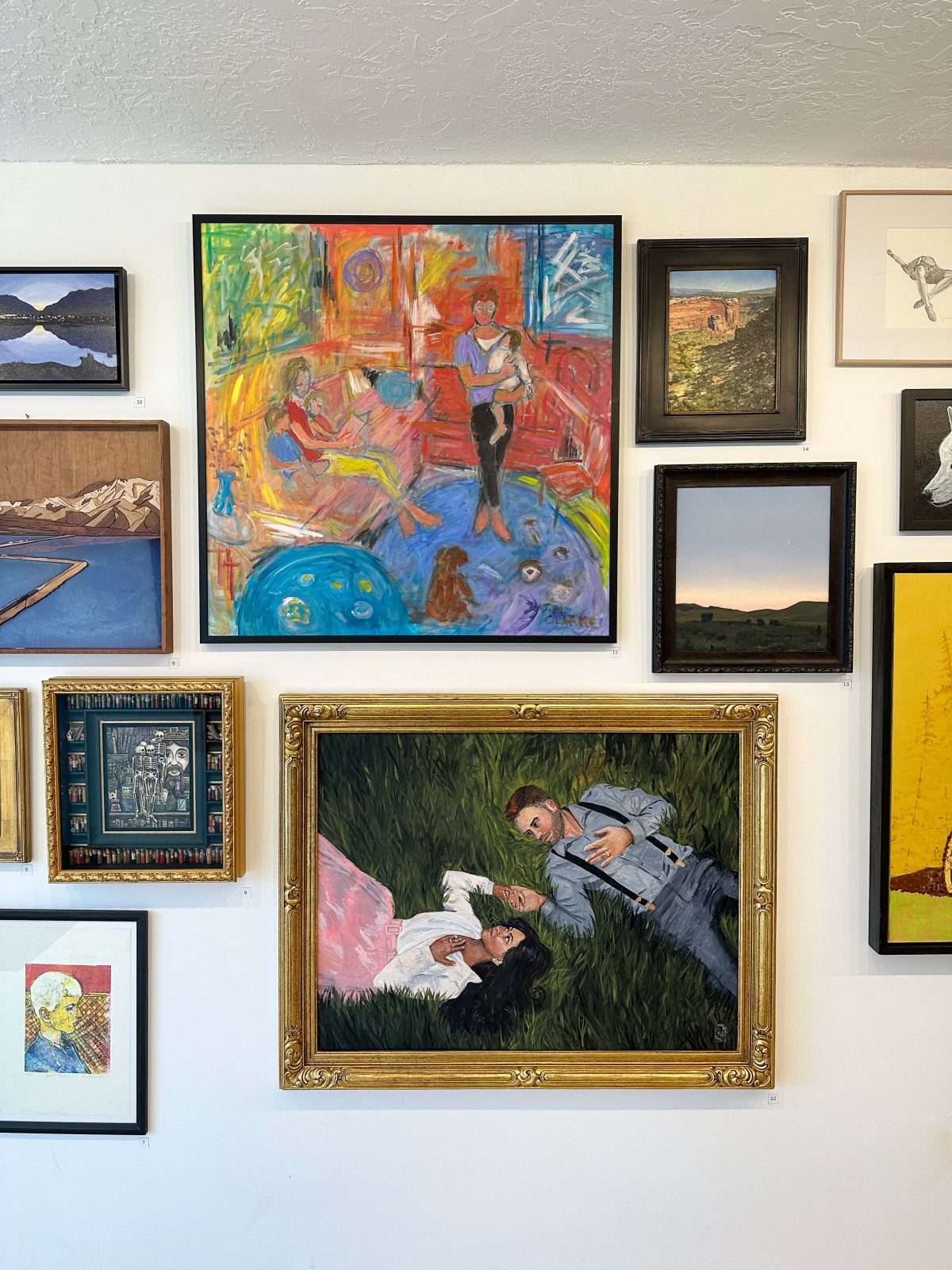 A close-up of a gallery wall showcasing a vivid abstract painting, a large framed painting of a man and woman lying in grass, and various smaller framed works including landscapes and portraits.