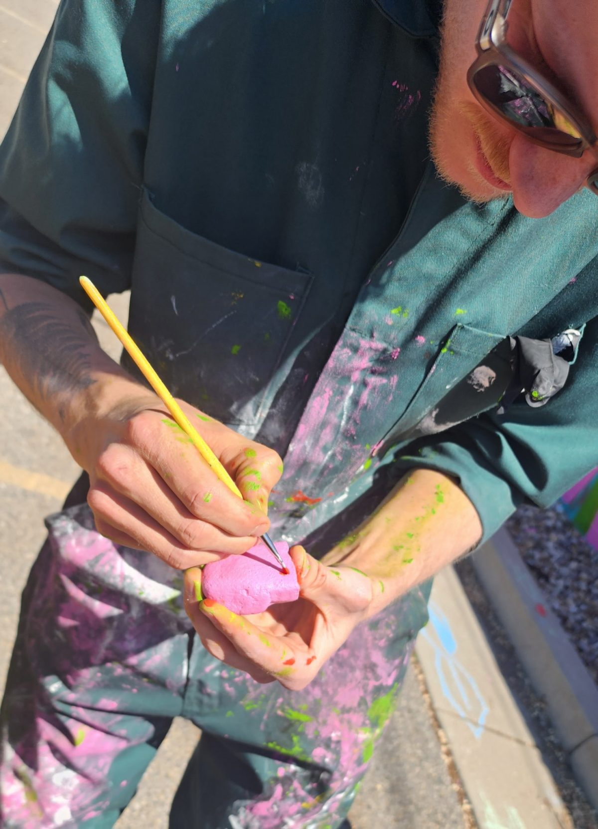 Close-up of an artist in a green jumpsuit holding and carefully painting a small pink object with a fine brush.
