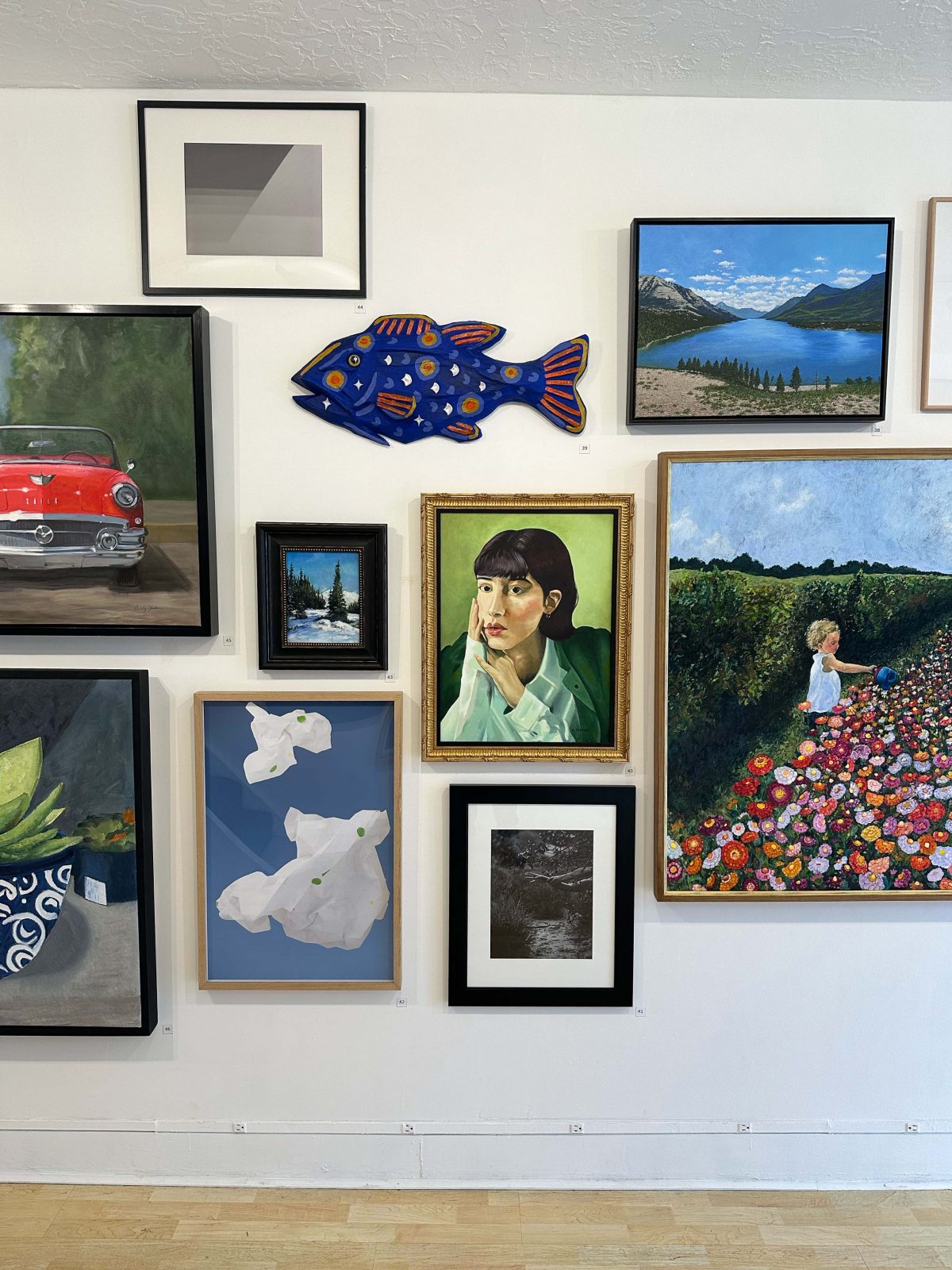 A section of gallery wall with an eclectic mix of artwork including a bright blue sculptural fish, a woman’s portrait, abstract paper-like forms, a vibrant flower garden scene with a child, and various landscape paintings.