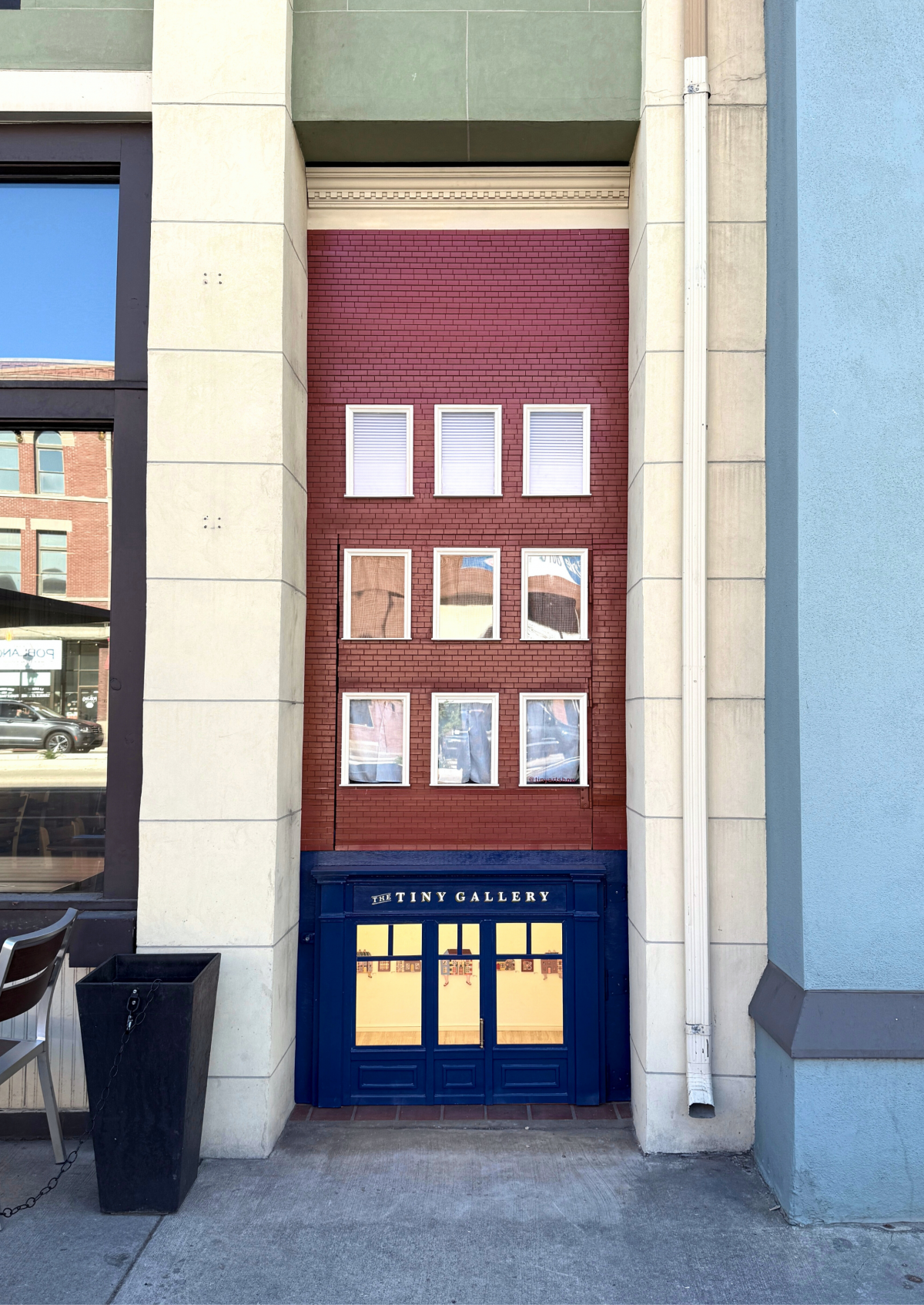 Full street view of The Tiny Gallery nestled between two storefronts, with its dollhouse-like facade mimicking the surrounding architecture.
