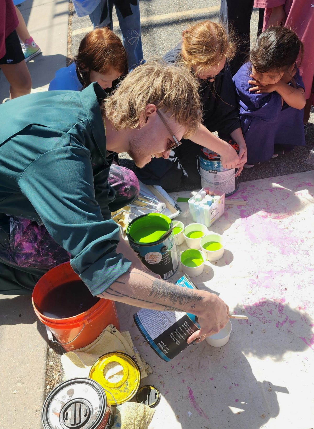 Artist kneels on pavement while pouring bright green paint from a can into smaller containers surrounded by art supplies and children.