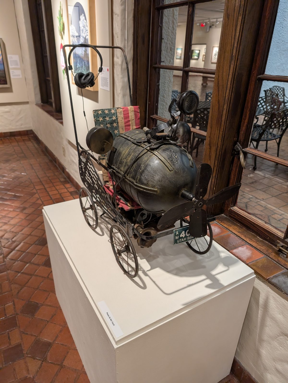 A metal contraption with a propeller, wheels, vintage radio headset, American flag fabric, and a small Utah license plate, displayed on a white pedestal.