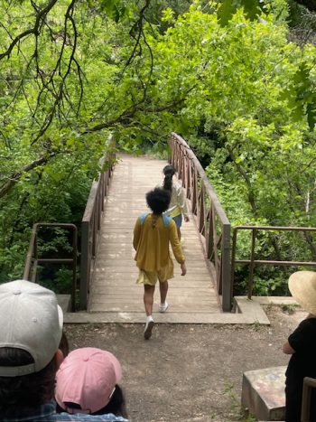 Audience members follow dancers in yellow and white costumes across a wooden bridge surrounded by green foliage at the Miller Bird Sanctuary.