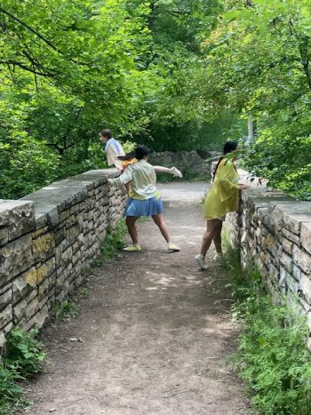 Three dancers in pastel costumes stretch and lean along a stone bridge amid dense forest, performing site-specific choreography.