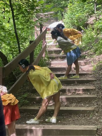 Three dancers in yellow and blue costumes lean forward while wearing headphones, performing on a rustic staircase in the forest.