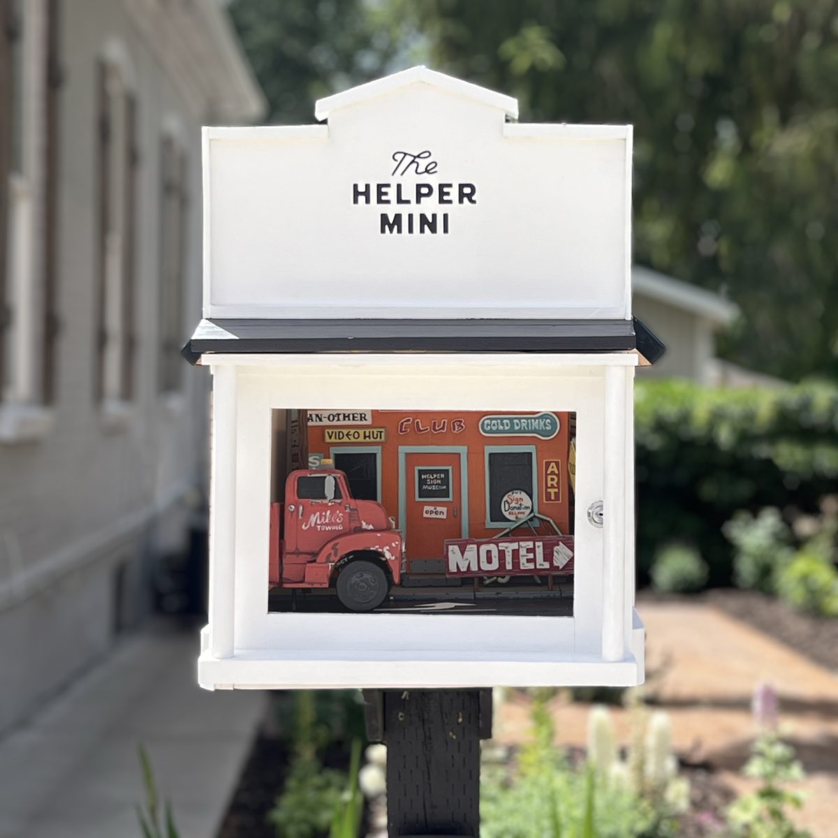 White miniature gallery box with “The Helper Mini” sign, displaying a detailed model of a vintage red truck parked in front of colorful storefronts with motel and art signage.