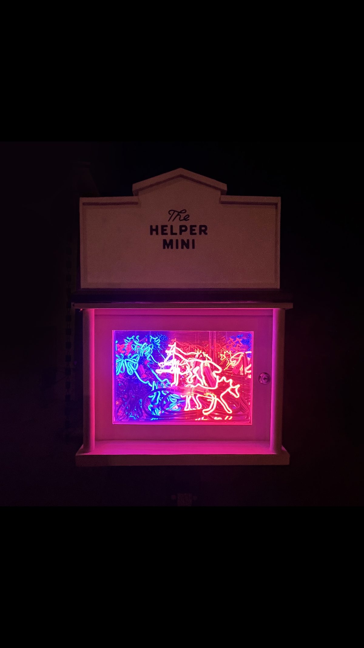 White miniature gallery box with “The Helper Mini” sign, glowing at night with layered neon lights in red, pink, and blue, forming stylized figures and shapes.