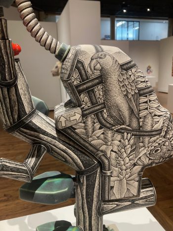 Close-up of a ceramic sculpture showing a parrot among leaves and pipes, its surface covered with finely drawn black-and-white imagery of nature and machinery.
