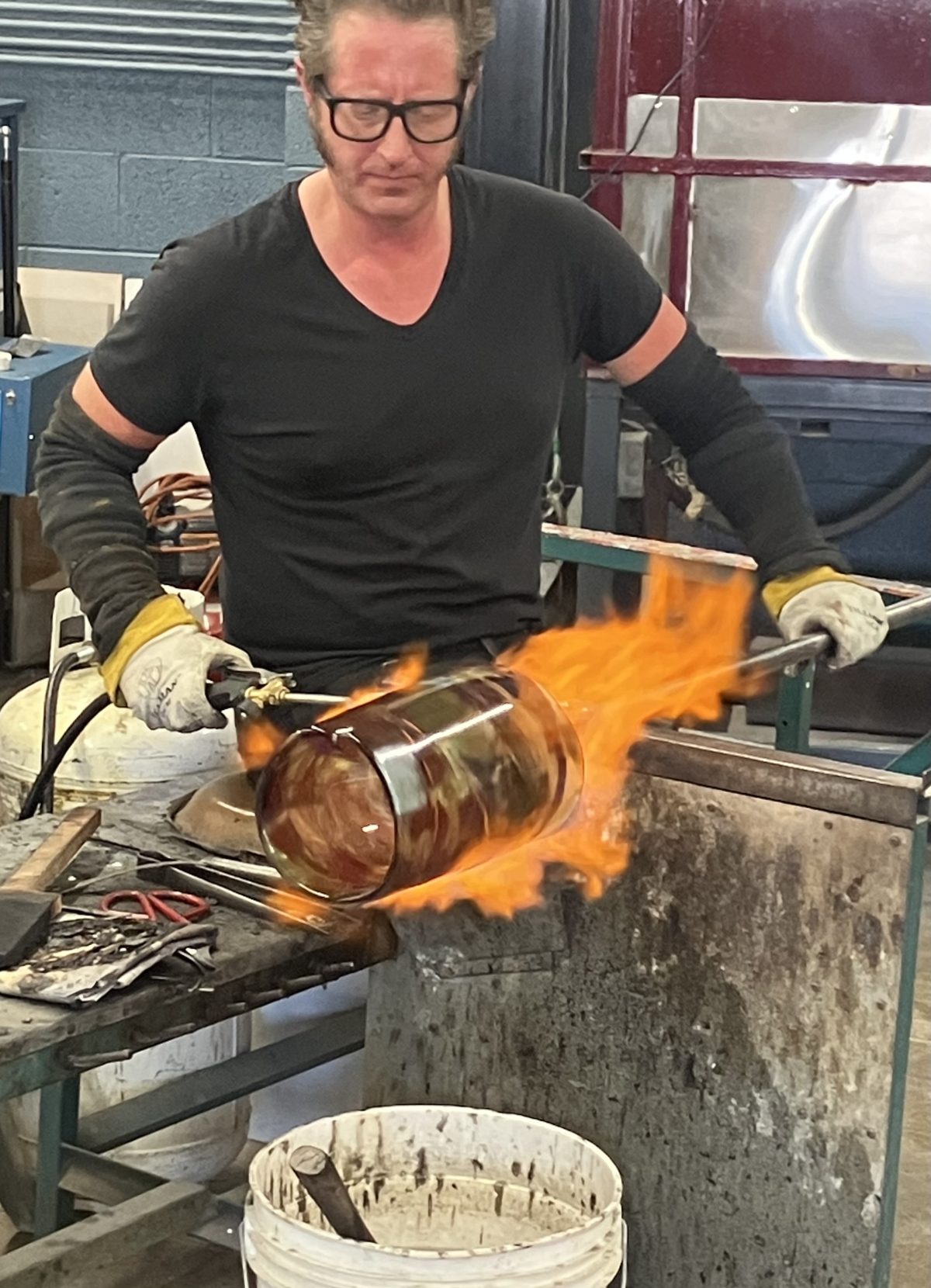 Glassblower Andrew Schafer shaping a molten vessel as flames engulf the surface during reheating.”