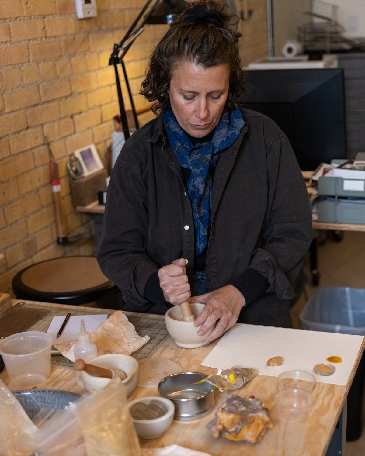 Elpitha Tsoutsounakis using a mortar and pestle to grind natural pigments at her studio table, surrounded by tools and materials.