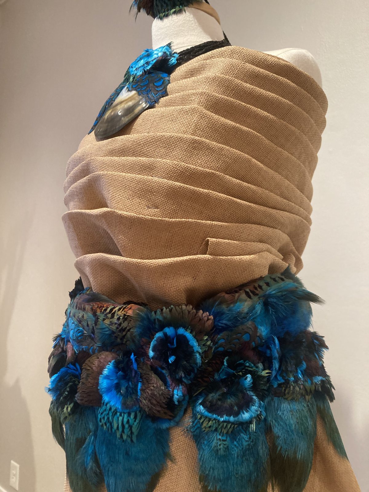 Close-up of a garment made from folded tapa cloth with a belt of blue and green iridescent feathers.