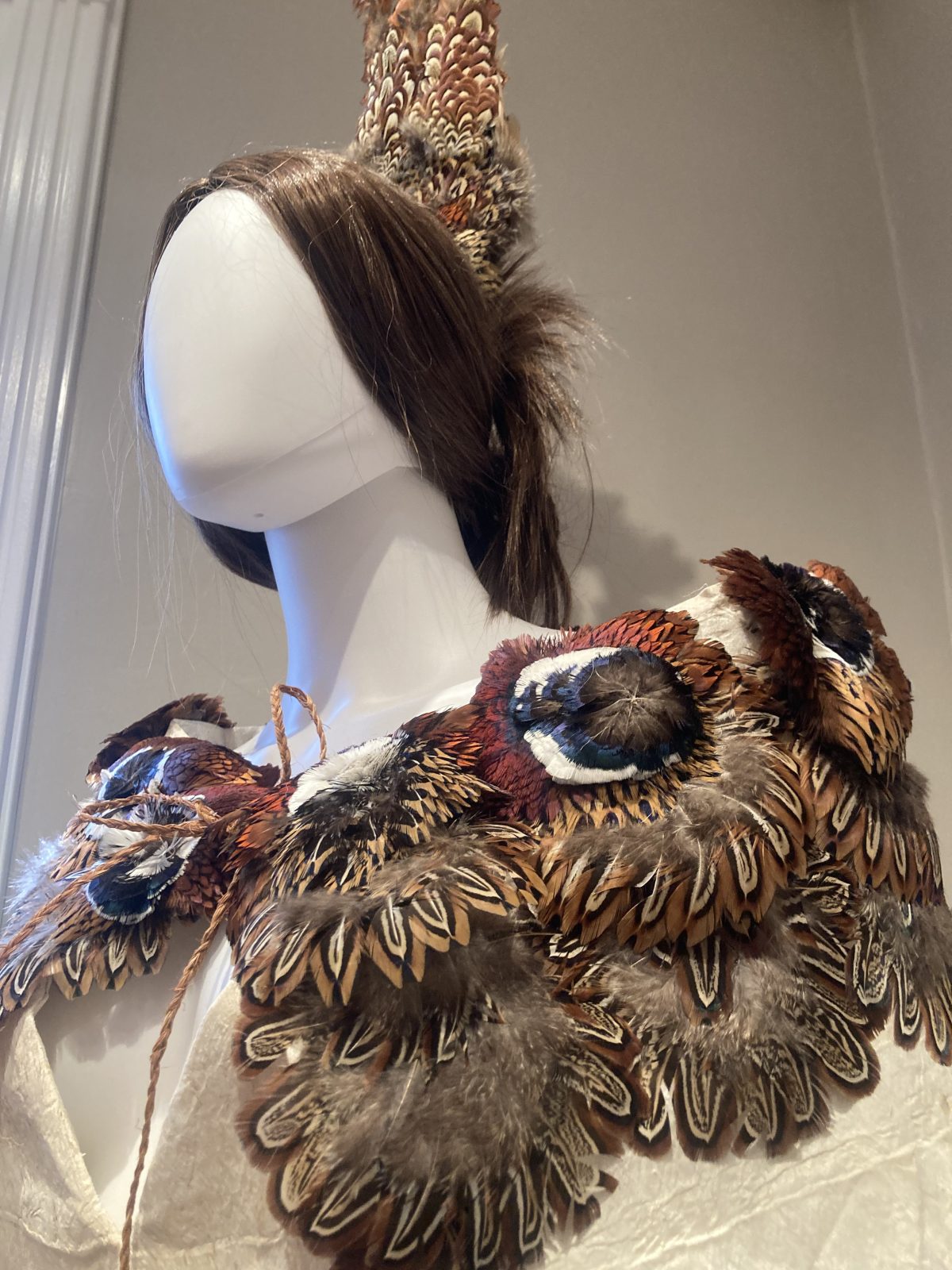 Close-up of a mannequin wearing a collar made of layered brown and patterned feathers with a tall feather headpiece.