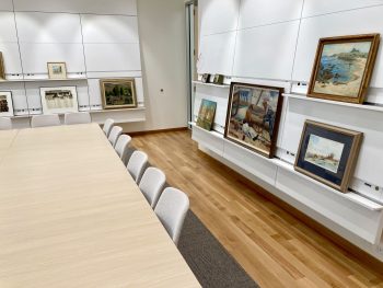 Museum study room with paintings displayed on wall-mounted racks beside a long table and chairs.