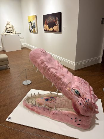 A monumental pink felt crocodile sculpture rests on the gallery floor with its jaws open wide. The oversized tongue contains embroidered imagery, while paintings and another soft sculpture are visible in the background.