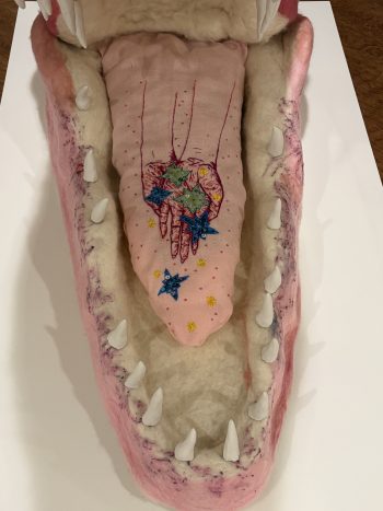 Close-up looking into the open mouth of a large pink felt crocodile sculpture. The soft pink tongue features embroidered hands surrounded by blue stars and small yellow flowers, framed by white felt teeth.