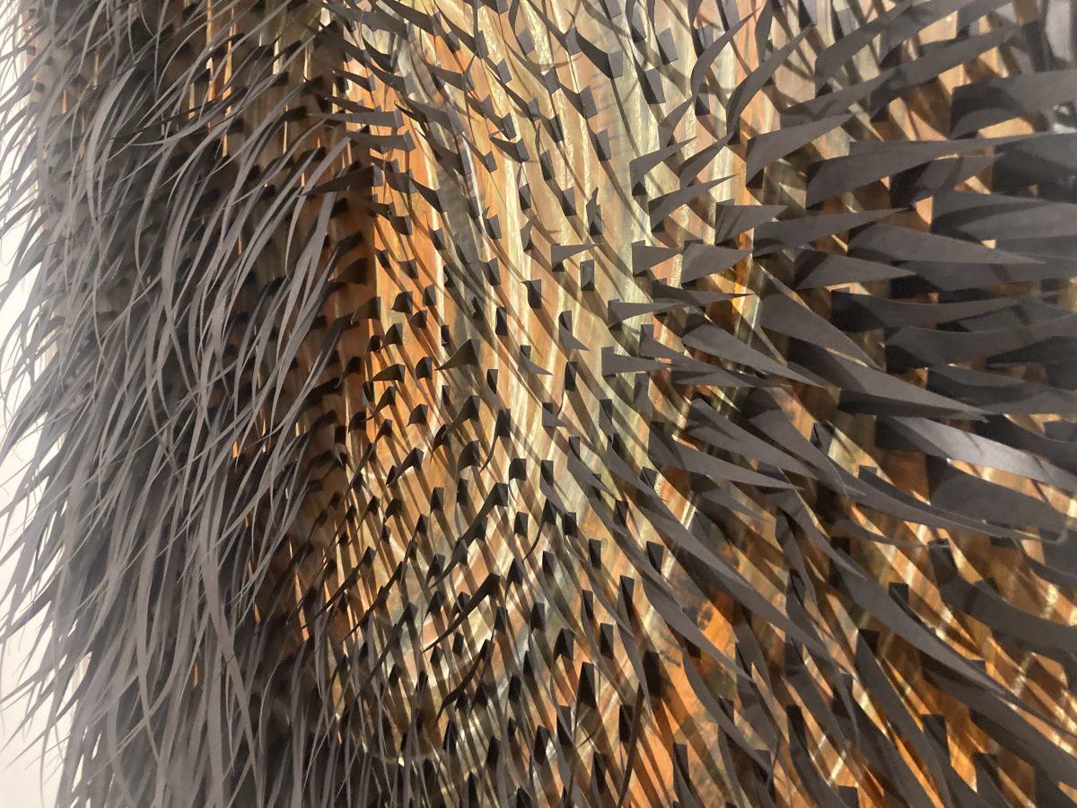 Close-up of a large mixed media paper work by Lenka Konopasek. Hundreds of narrow black paper strips project outward from a layered surface of warm gold, bronze, and ochre tones, creating a dense, tactile texture.